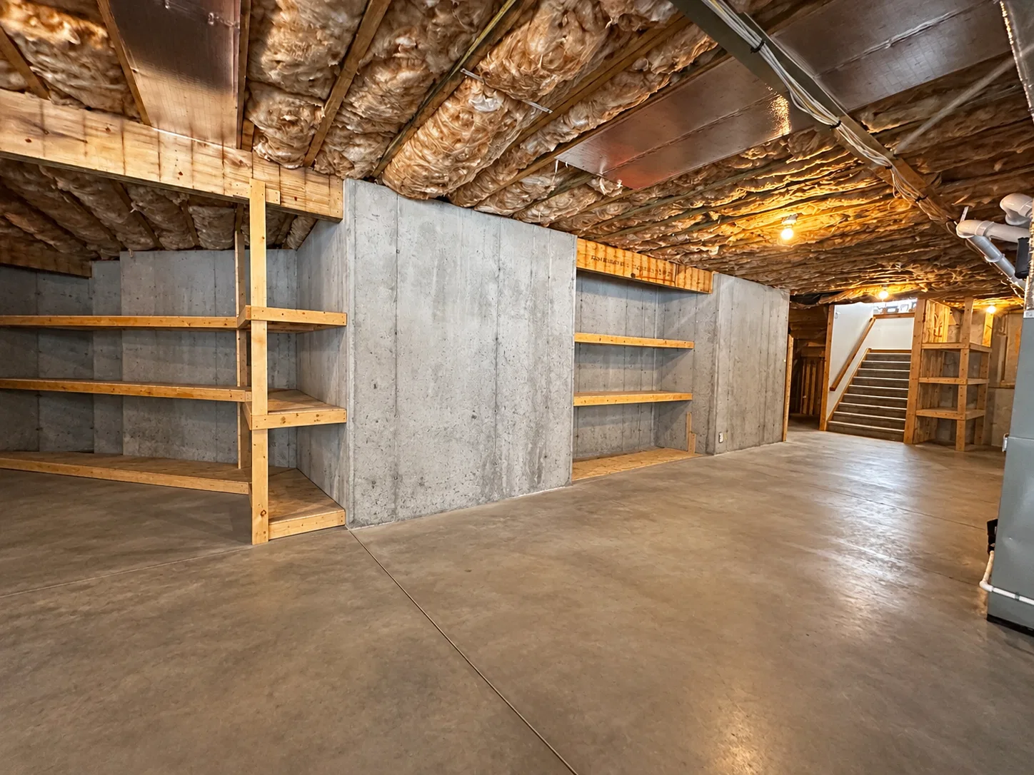 Before finishing: wood storage shelves against concrete basement walls, with exposed ceiling insulation and stairs in the background.