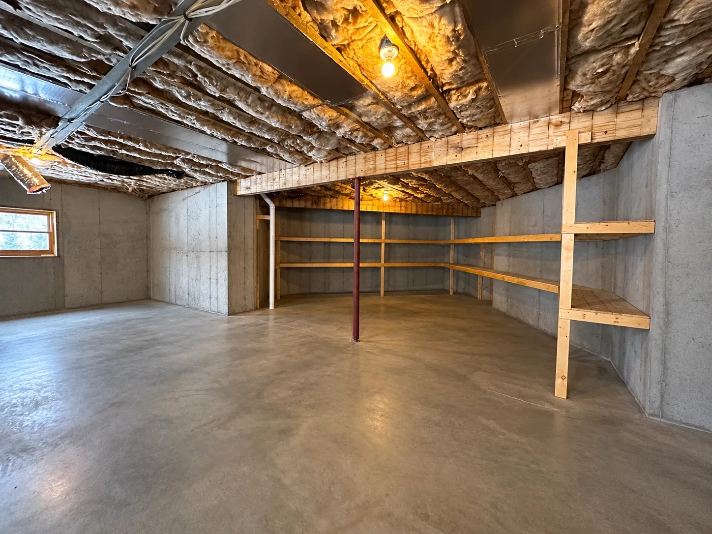 Before finishing: basement storage corner with bare wood shelves, concrete walls, a window and a steel support post beneath exposed insulation.