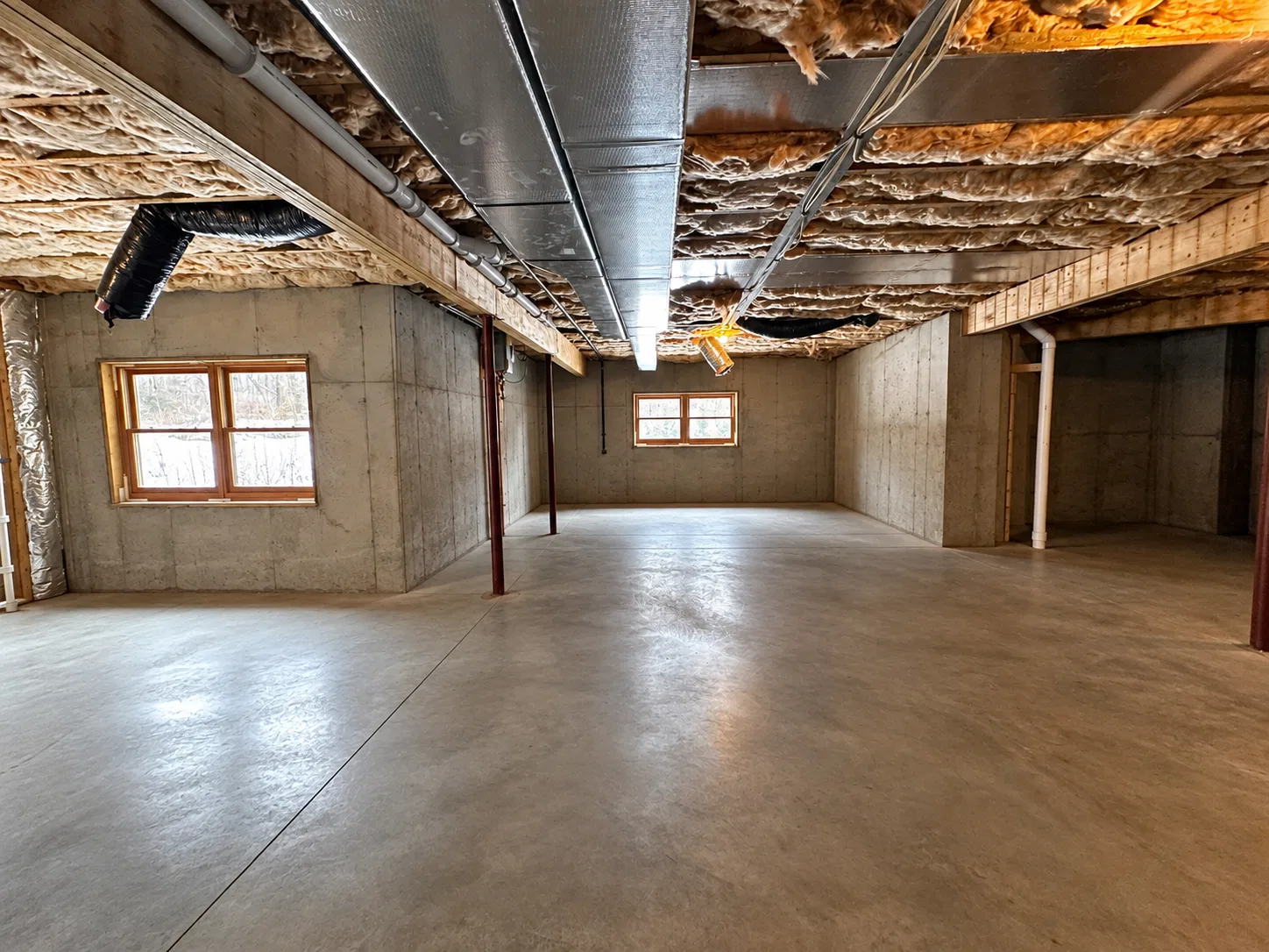 Before finishing: long view of a concrete basement floor beneath metal ductwork, with windows, steel posts and an unfinished storage recess.
