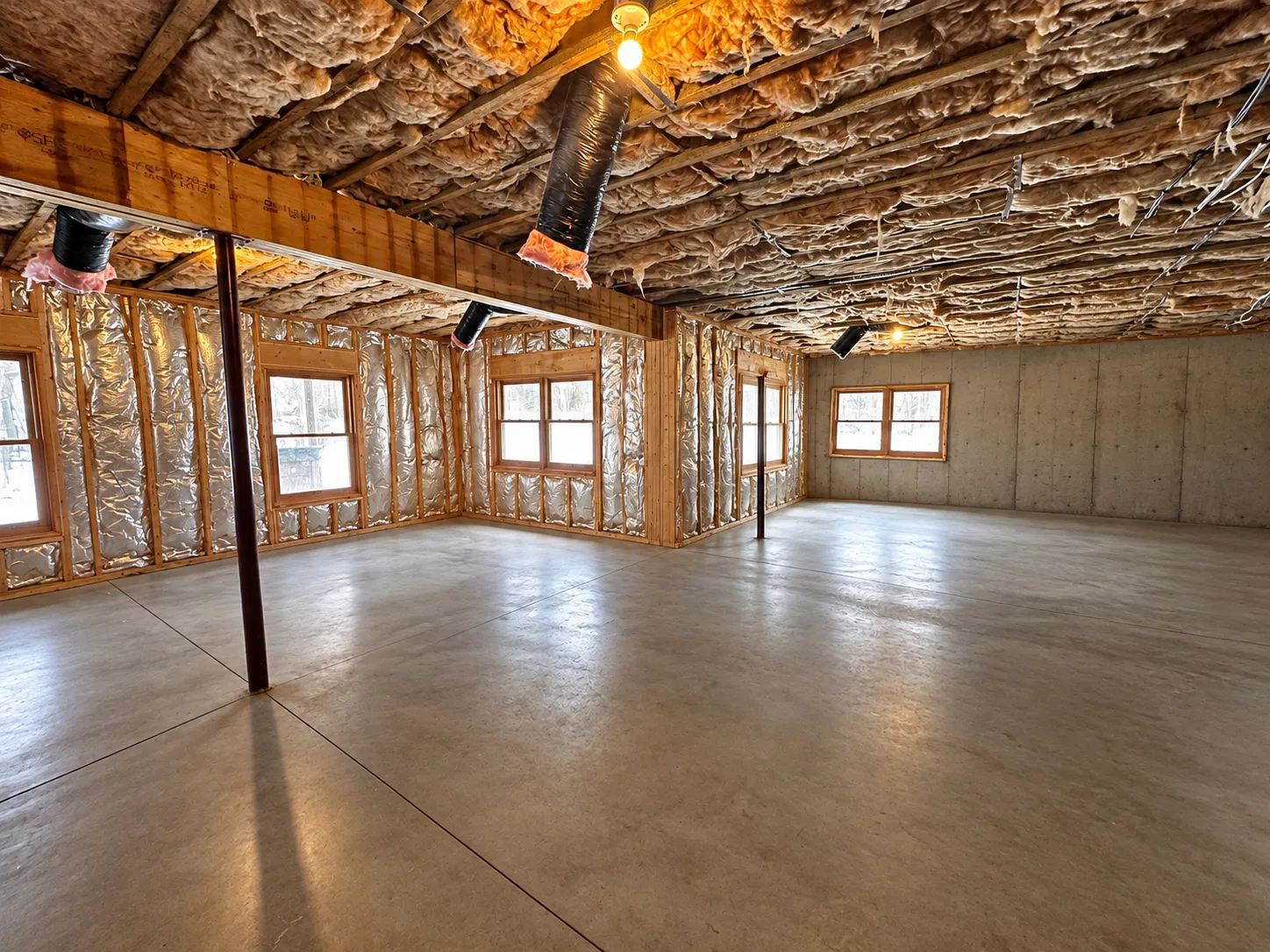 Before finishing: open basement corner with several windows, exposed insulated wall framing, steel support posts and bare concrete flooring.