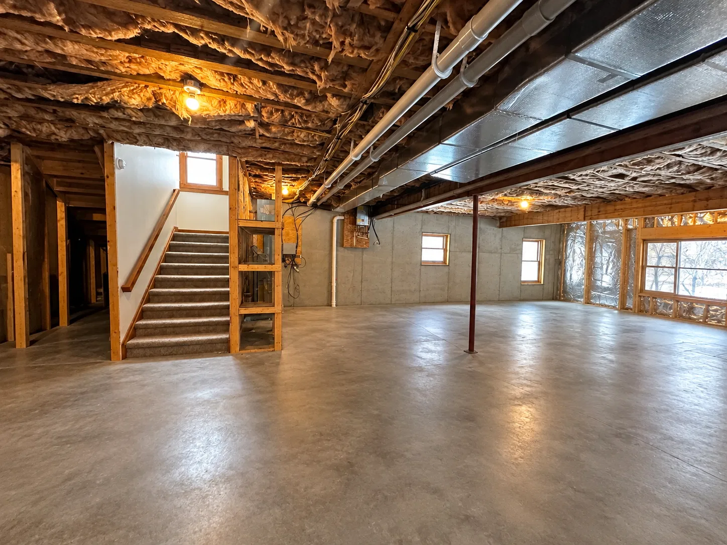 Before finishing: carpeted basement stairs beside open wood wall framing, overhead ductwork, a steel post and bare concrete flooring.