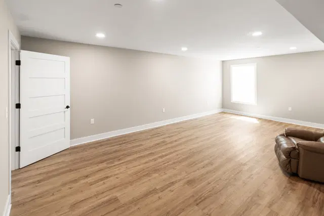 Open basement room with wood-look flooring, gray walls, white doors and a sunlit window.