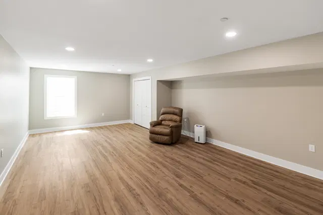 Wide basement room with wood-look flooring, a window and brown recliner beneath a boxed ceiling soffit.