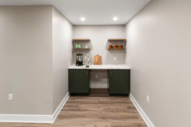 Recessed basement beverage nook with dark cabinets, a white counter, small refrigerator and floating shelves.