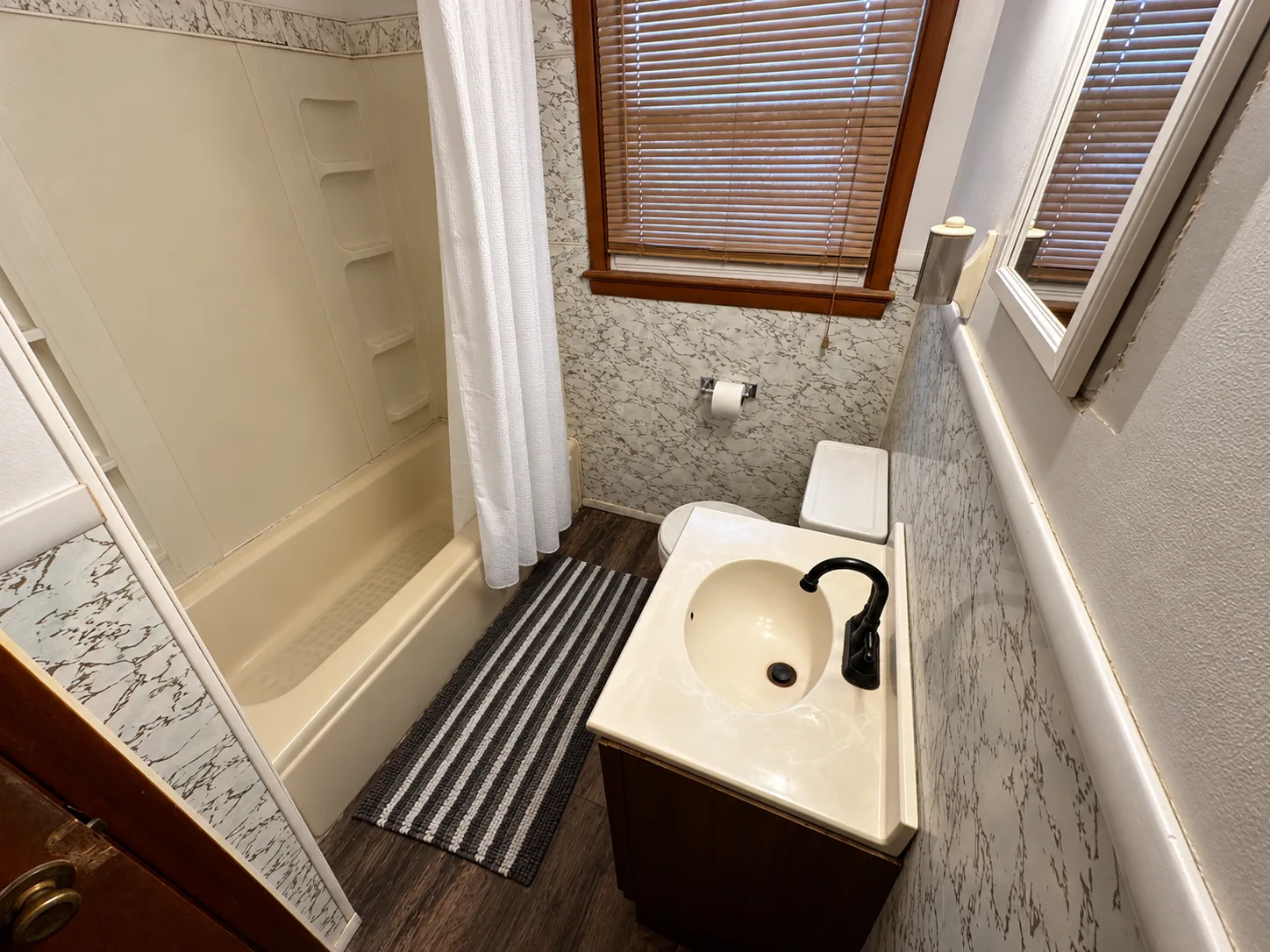 Before remodeling: Middletown bathroom with a cream tub-and-shower combination, white shower curtain, wood vanity, toilet, marble-pattern wall panels and a wood-trimmed window with blinds.