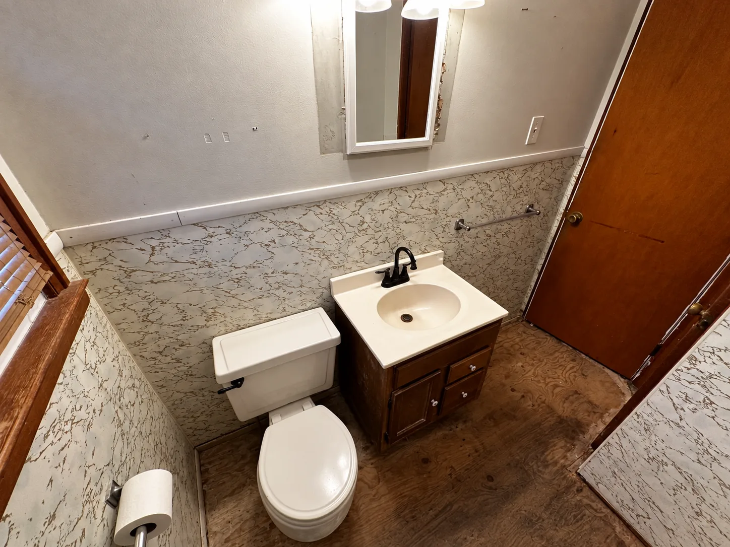 Before remodeling: small wood vanity with a cream sink and black faucet beside a white toilet, marble-pattern lower wall panels and exposed wood subfloor.