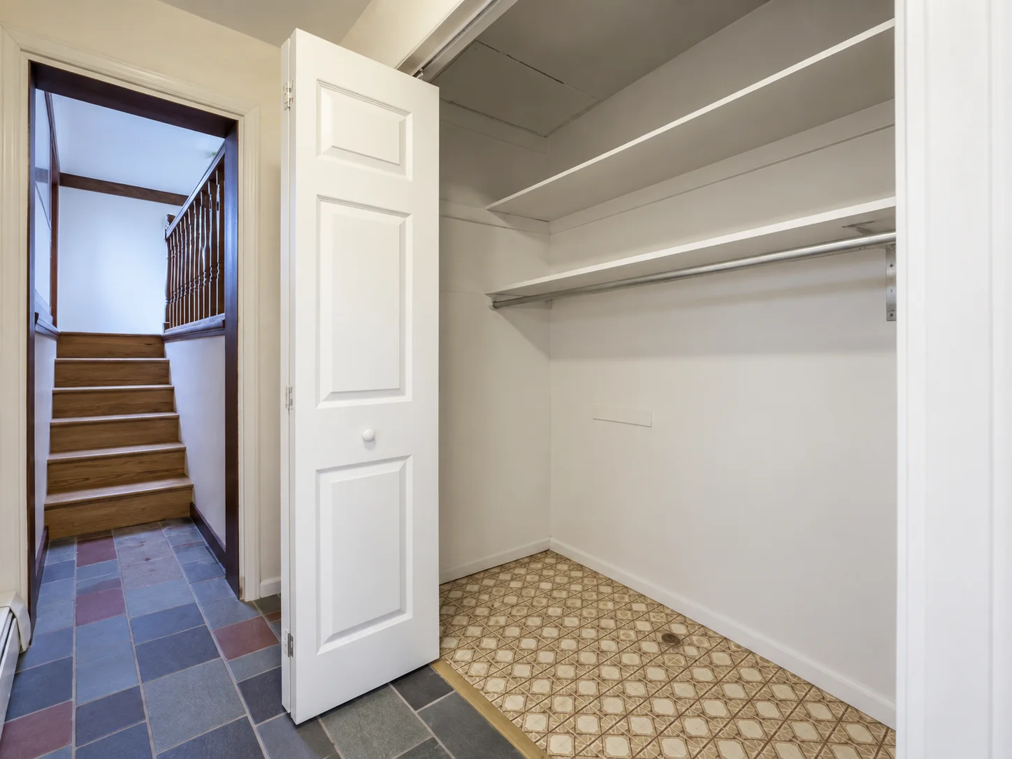 Before remodeling: empty closet in Palmyra with white shelves, a hanging rail, folding doors and patterned flooring beside a tiled hallway and wooden stairs.