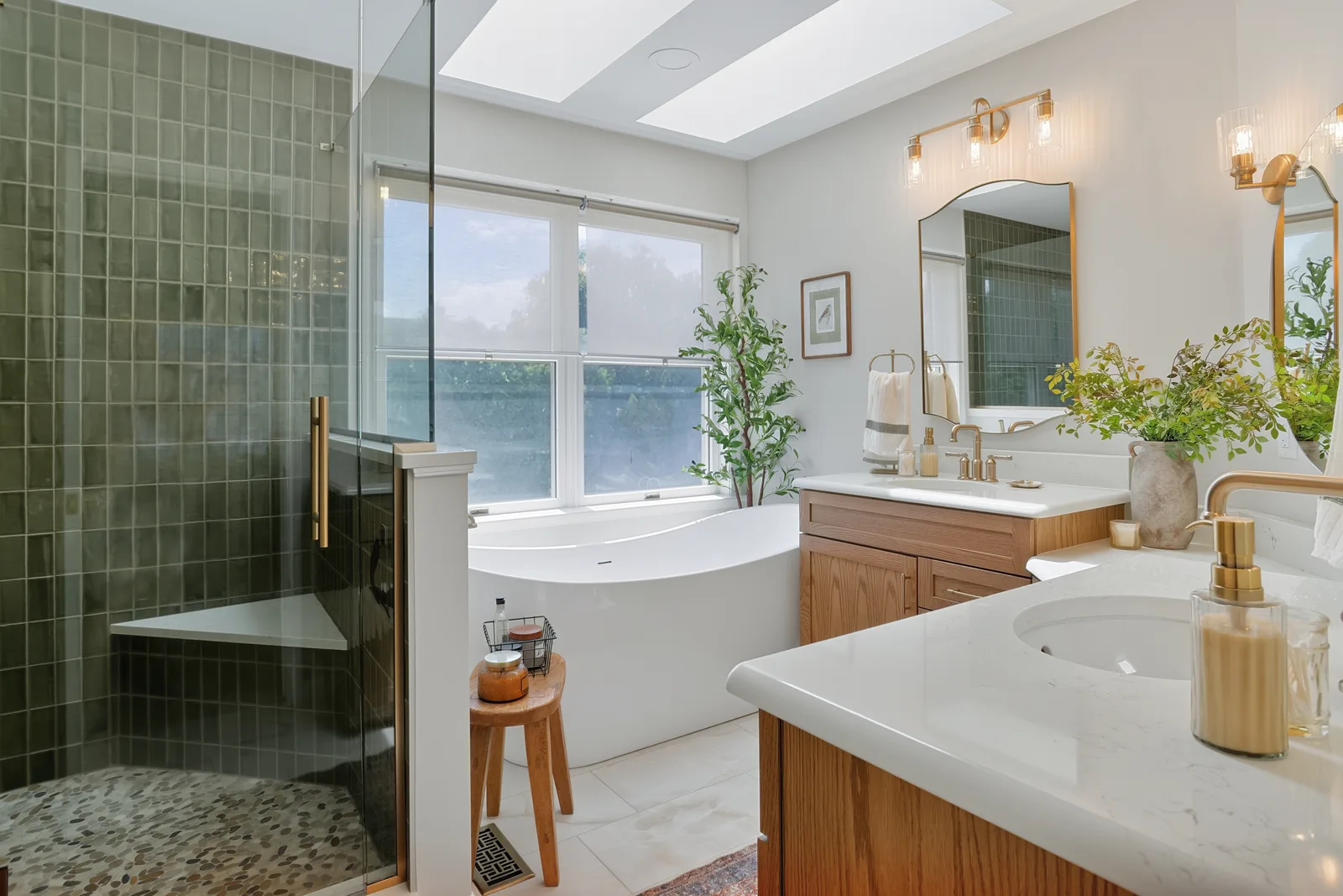 Bathroom with a green-tiled glass shower, freestanding tub, wood vanities and gold-tone fixtures.