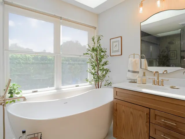 Freestanding bathtub beneath a large window beside a wood vanity and arched mirror.