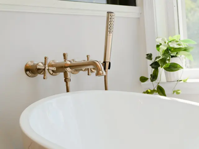 White freestanding bathtub with a gold-tone wall-mounted filler beneath a window.