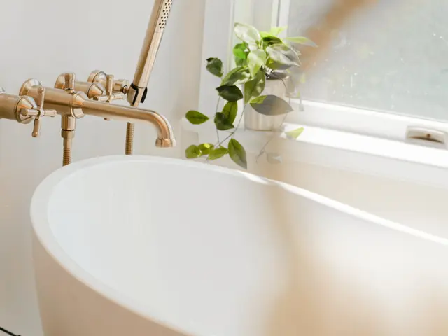 Wall-mounted gold-tone bathtub filler above a white tub beside a sunlit window.