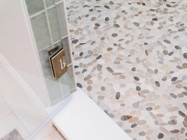 Glass shower hinge beside the speckled mosaic floor and green tile walls.