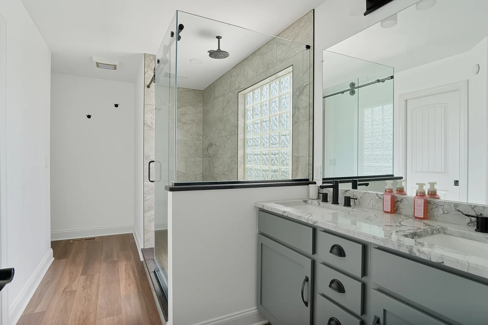 Finished bathroom overview with a glass-enclosed shower, gray vanity, black-framed mirror, marble-look countertop and wood-look floor.
