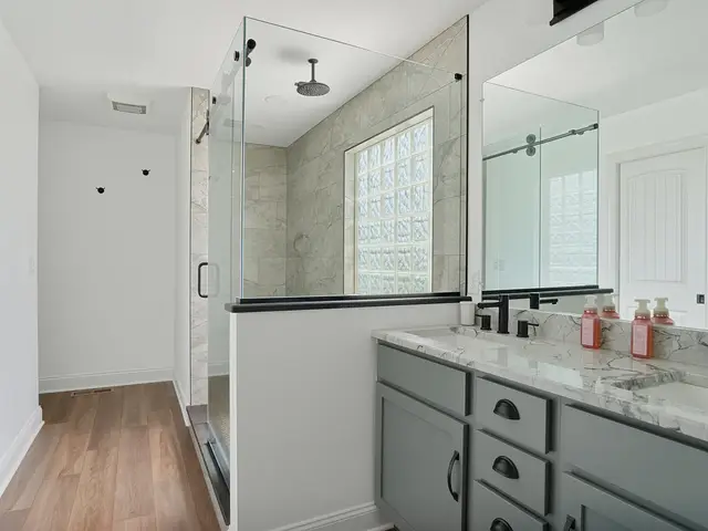 Finished bathroom overview with a glass-enclosed shower, gray vanity, black-framed mirror, marble-look countertop and wood-look floor.