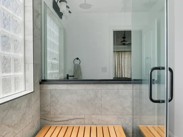 View down the glass shower showing gray wall tile, a glass-block window and a wood shower bench.