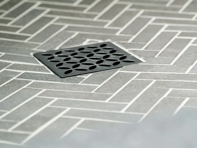 Square dark shower drain set into herringbone-pattern floor tile.