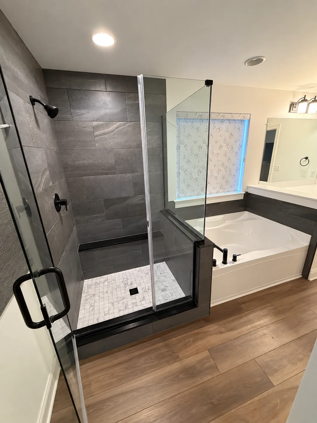 Before remodeling: glass-enclosed shower with dark rectangular wall tile and a light mosaic floor beside a built-in bathtub and shaded window.