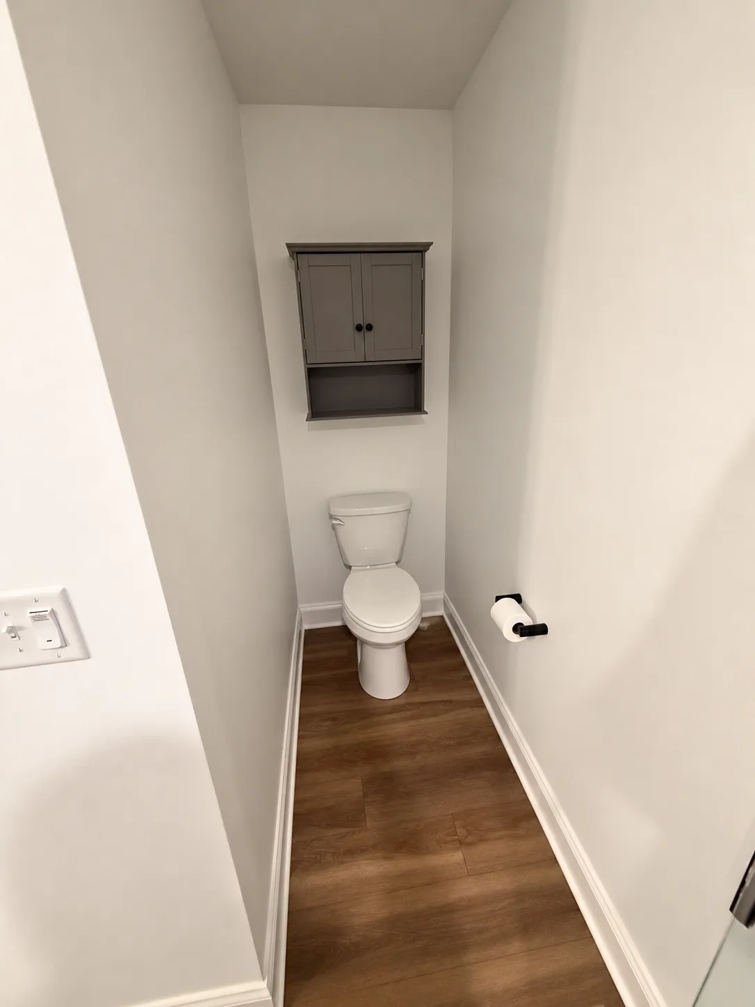 Before remodeling: separate toilet alcove with a gray wall cabinet, white walls and wood-look flooring.