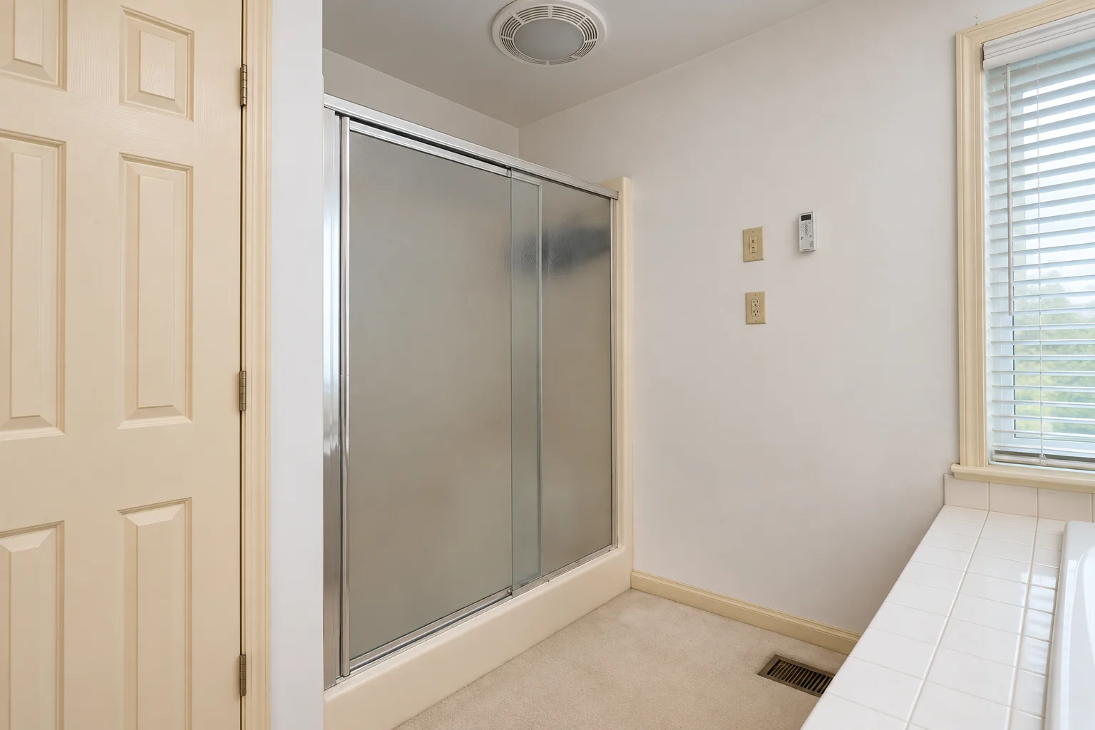 Before remodeling: framed shower enclosure with frosted sliding doors beside the built-in tub and beige carpet.
