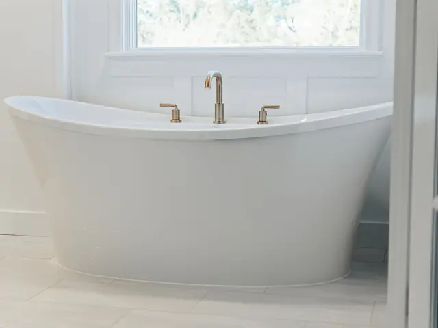 Freestanding tub with a brushed-gold faucet beneath a window and white paneled walls.
