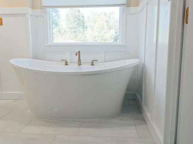 Freestanding tub centered under a window with white wainscot paneling and light tile flooring.