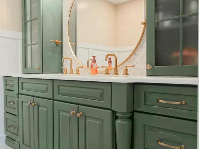 Sage green vanity with a white countertop, gold-tone faucets and a round mirror.