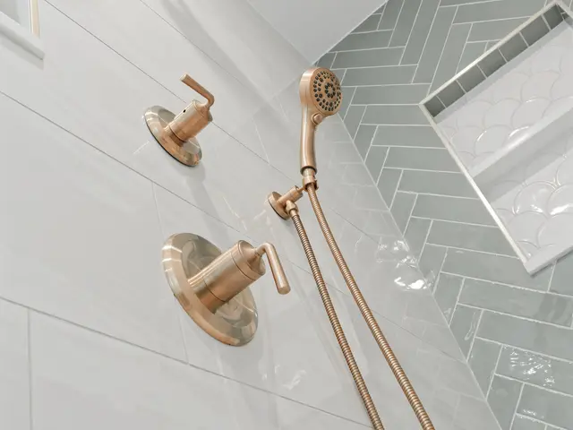 Gold-tone shower valve and handheld spray beside a green herringbone tile niche.