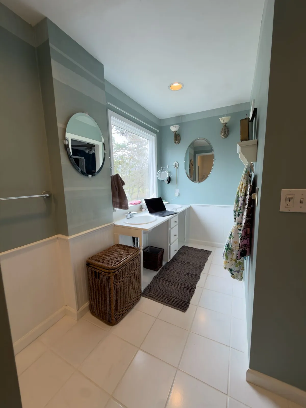 Before remodeling: narrow vanity with two sinks, round mirrors, a window and a wicker hamper above white tile flooring.