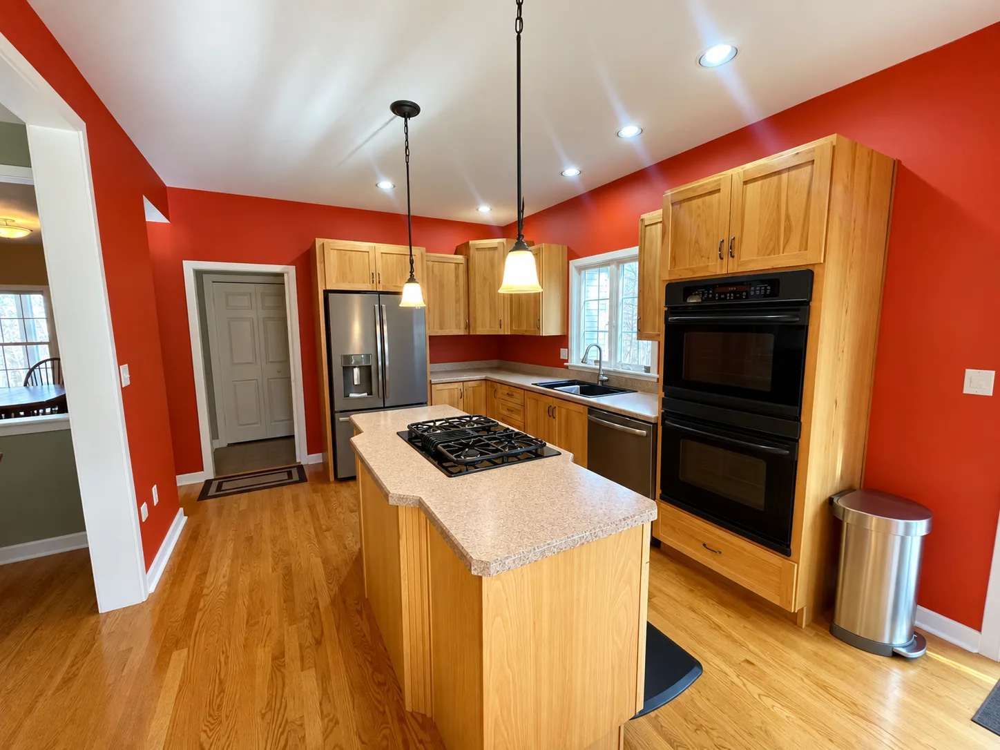 Before remodeling: wood kitchen island with a gas cooktop, stacked black ovens, stainless refrigerator and sink beneath a window, surrounded by red walls.