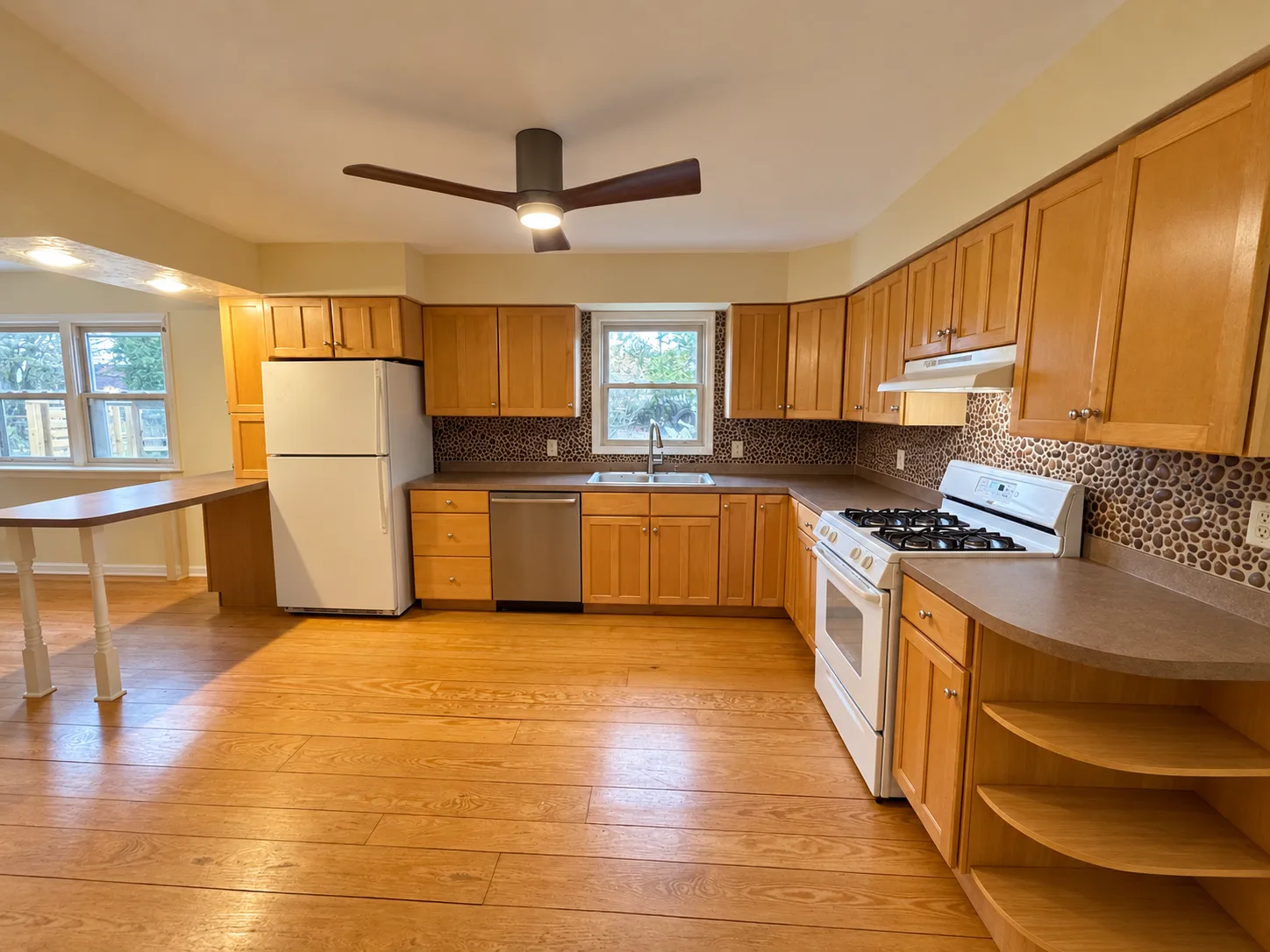 Before remodeling: wood kitchen cabinets surrounding a white refrigerator, sink, stainless dishwasher and white gas range beneath a pebble-pattern backsplash.
