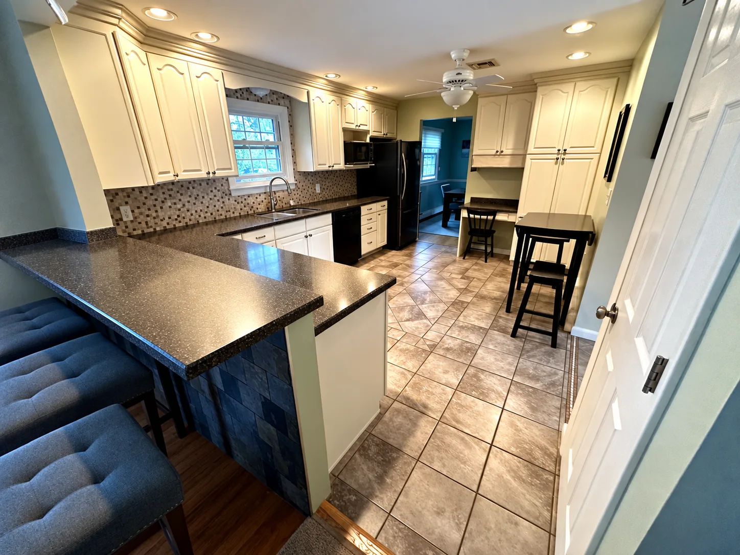 Before remodeling: Camp Hill kitchen with cream raised-panel cabinetry, dark countertops, a mosaic backsplash, black refrigerator, patterned floor tile and a peninsula beside upholstered stools.