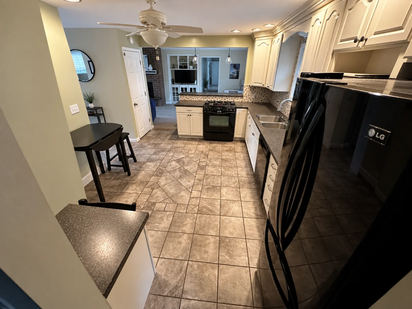 Before remodeling: kitchen with a black range facing the adjoining living area, cream cabinets, a double sink, black refrigerator, square floor tile and a ceiling fan.