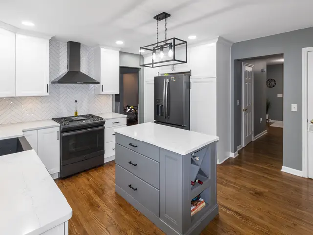View across the gray kitchen island showing drawers and wine storage, dark appliances, white cabinetry and warm-toned flooring.