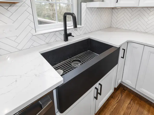 Close-up of a black apron-front kitchen sink and black faucet surrounded by light countertops, white cabinets and herringbone tile.