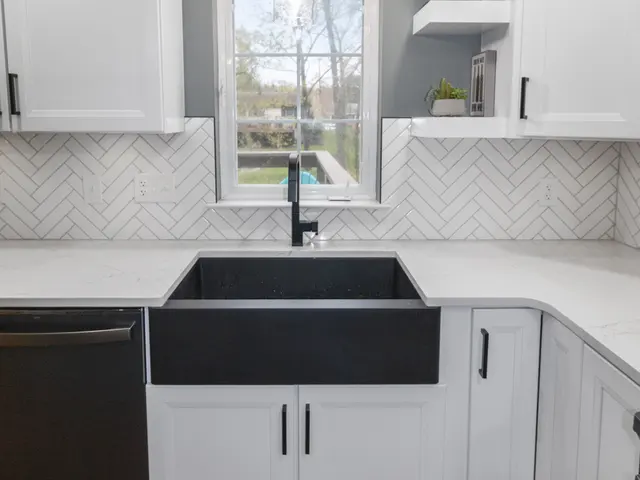 Black apron-front sink beneath a window and glass pendant, with white kitchen cabinets, floating corner shelves and a herringbone backsplash.