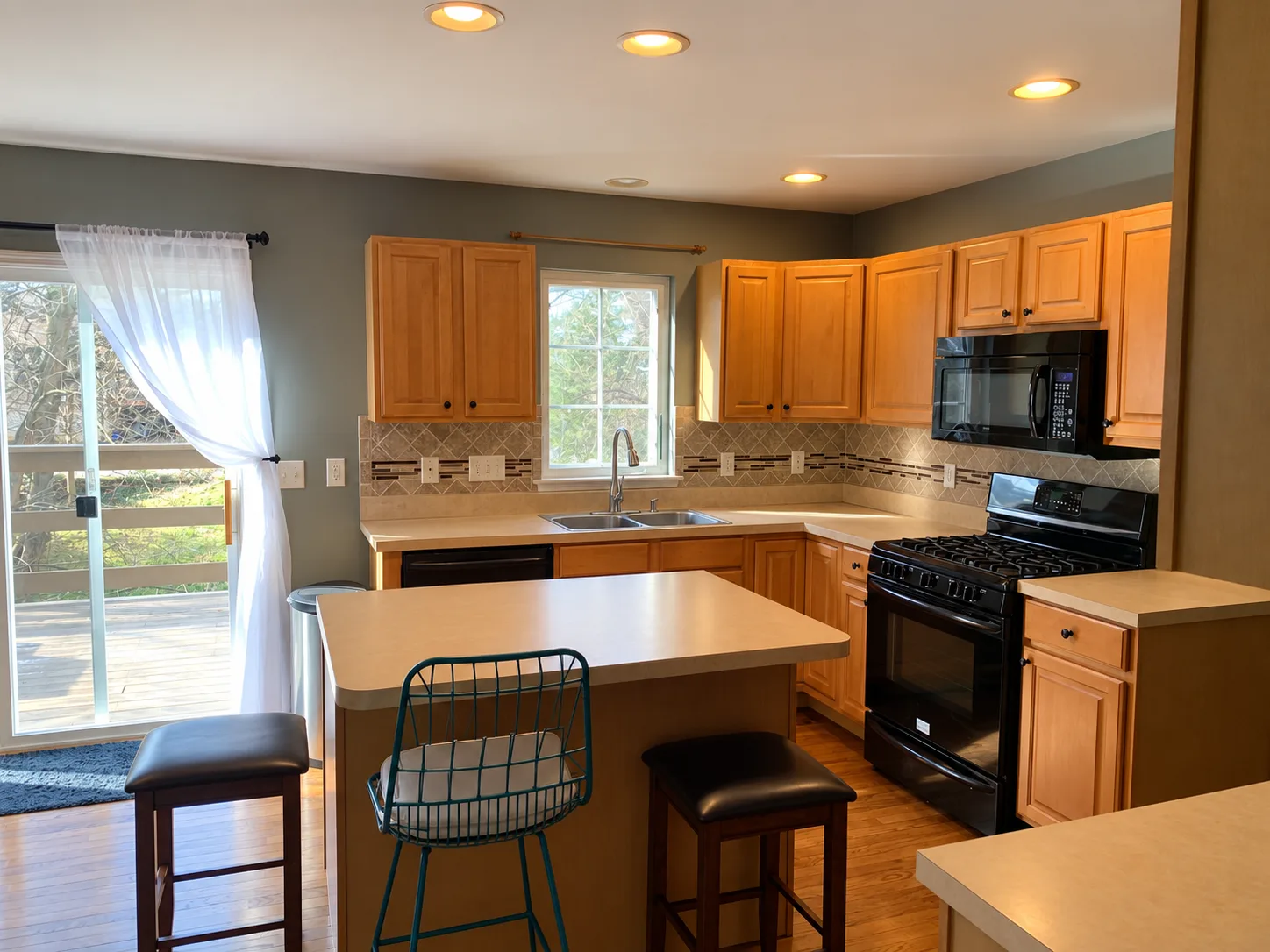 Before remodeling: wood kitchen cabinetry around a stainless sink beneath a window, with black appliances and stools beside the island near a sliding glass door.