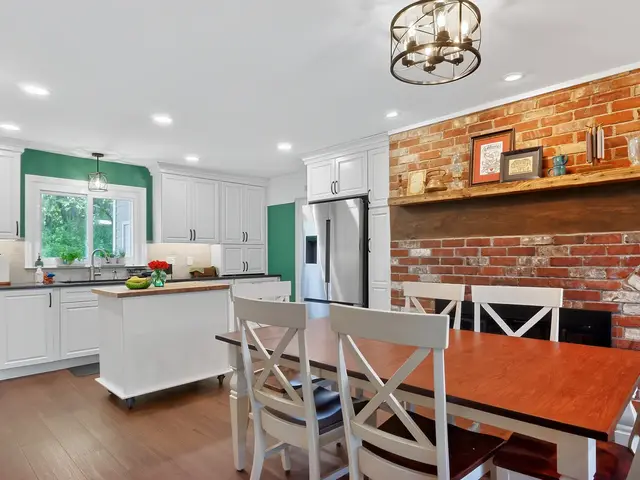 Wide view of the finished white kitchen with a wood-topped island, green window accents and a dining area beside a brick fireplace.