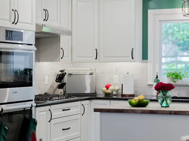 Wide kitchen view showing stacked wall ovens, the gas cooktop, white corner cabinetry, the island and sink beneath a window.
