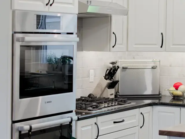 Vertical view of the kitchen cooking area with stacked stainless ovens, white cabinets and a gas cooktop beneath a vent hood.
