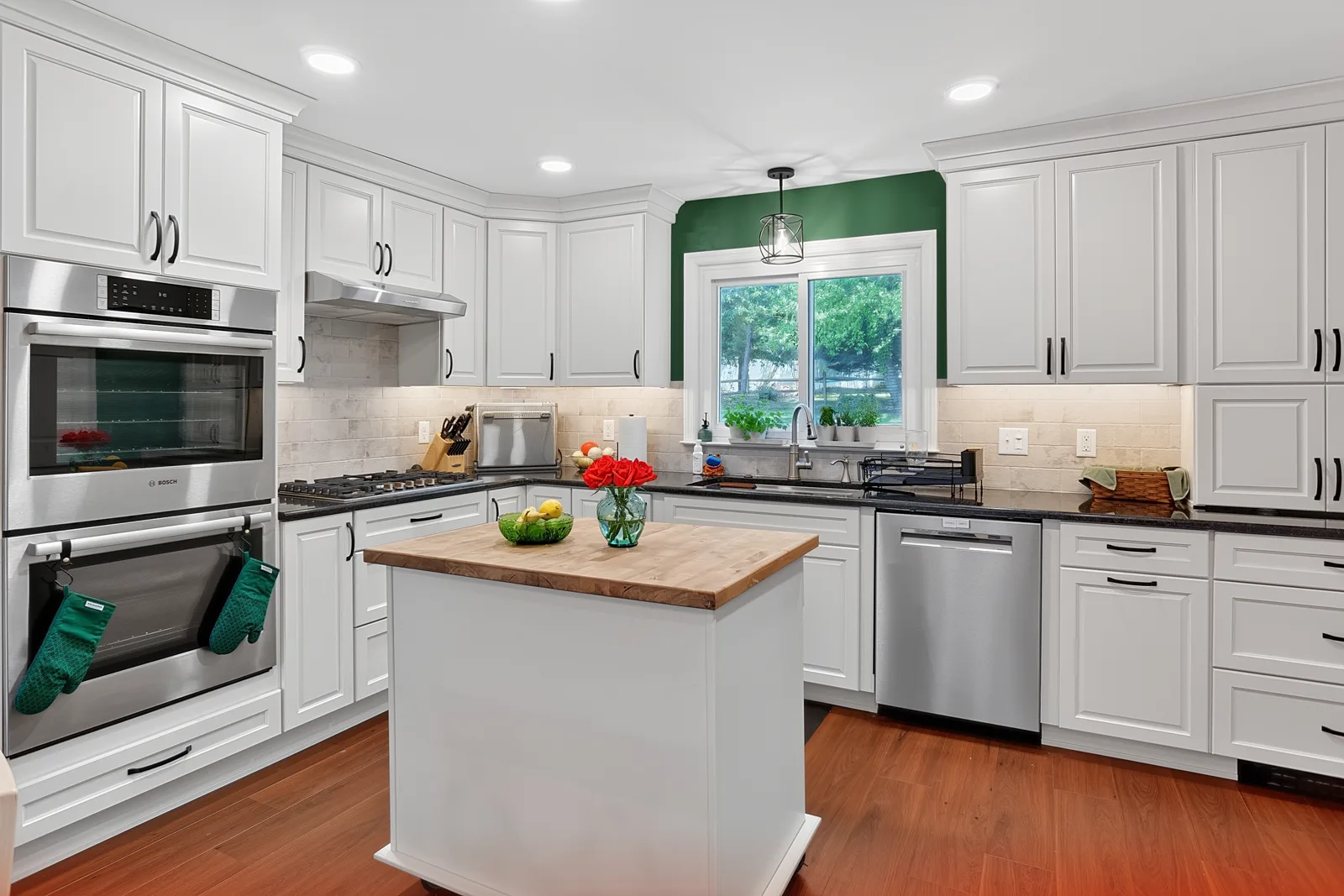 Overview of the finished white kitchen with a wood-topped island, stacked stainless ovens, gas cooktop, dark countertops and a green accent wall around the sink window.