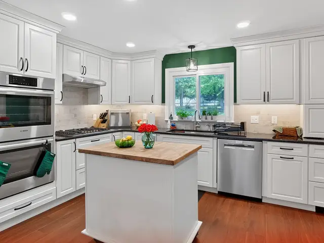 Overview of the finished white kitchen with a wood-topped island, stacked stainless ovens, gas cooktop, dark countertops and a green accent wall around the sink window.