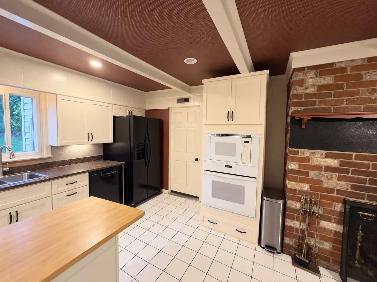 Before remodeling: kitchen with black refrigerator and dishwasher, a white microwave-and-oven cabinet, a wood-topped island and the adjoining brick fireplace.