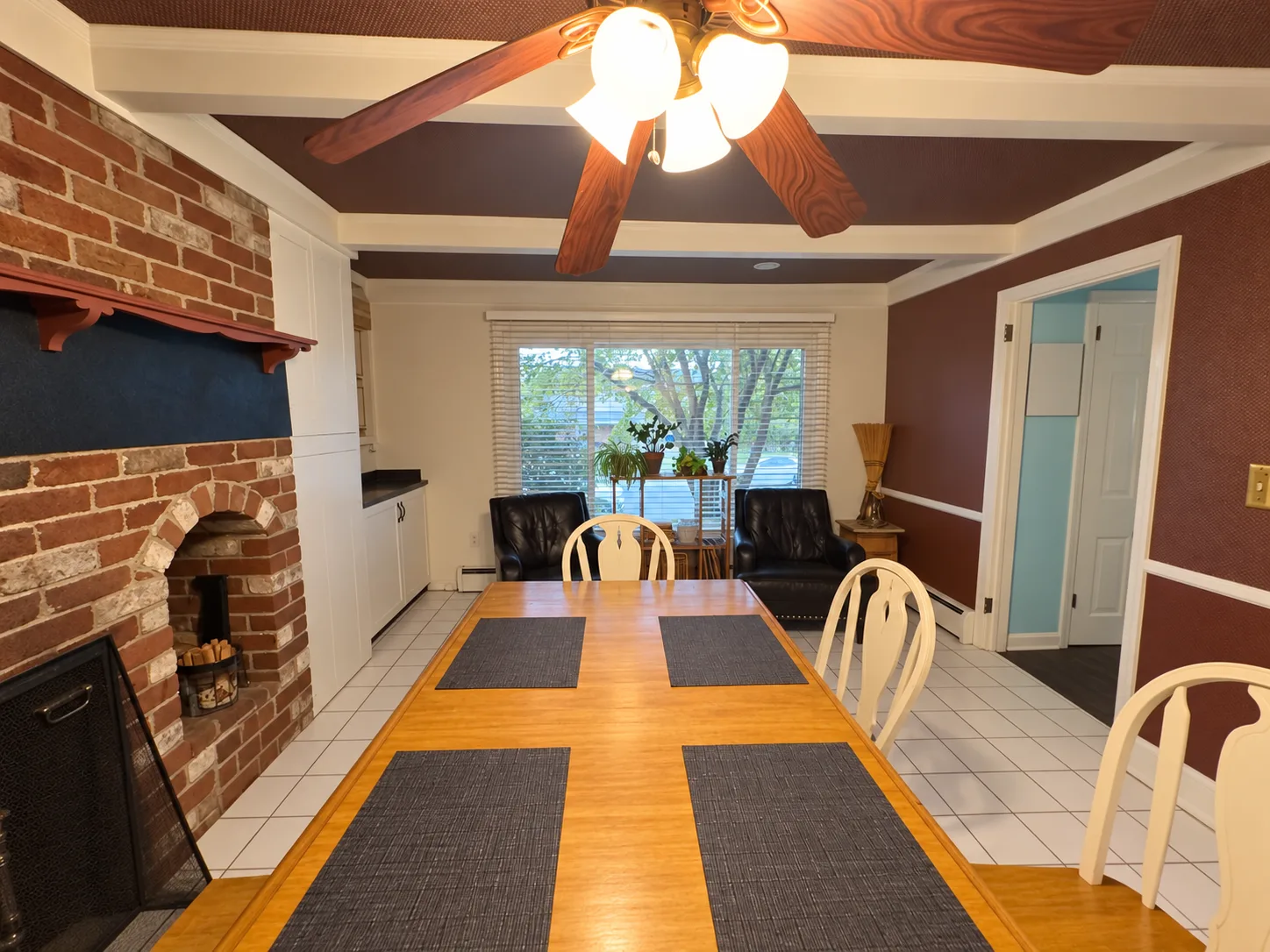 Before remodeling: dining area with a wood table, brick fireplace, white tile floor, window-side seating and a ceiling fan beneath white beams and a brown ceiling.