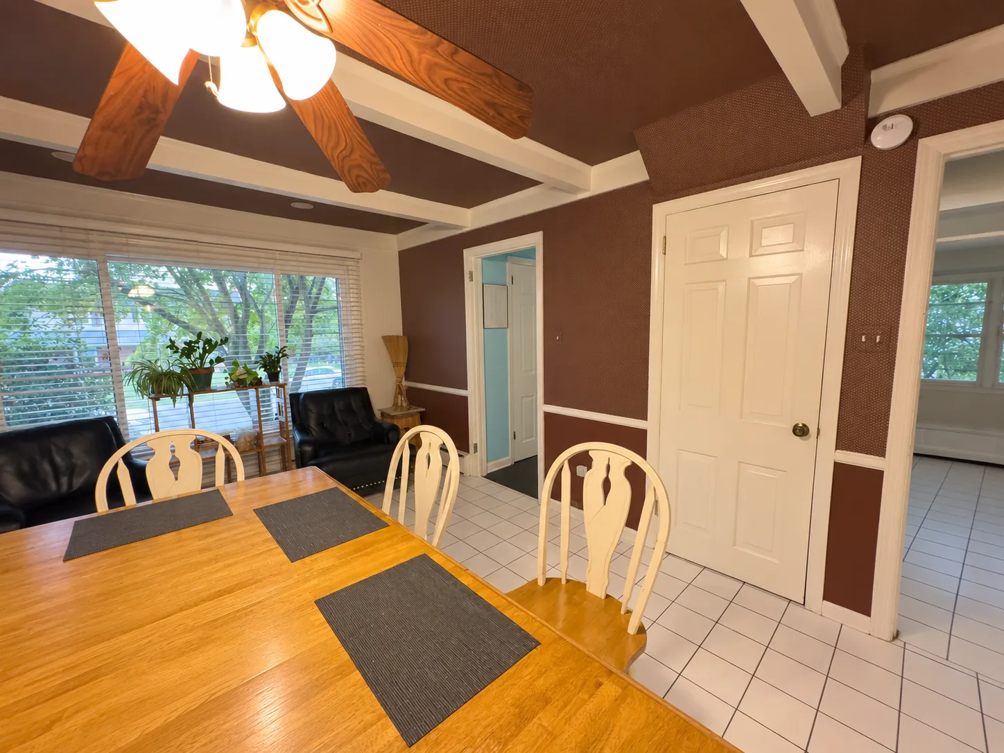Before remodeling: dining table and chairs beside large windows, brown patterned walls, white doorways and a white tile floor beneath a ceiling fan.