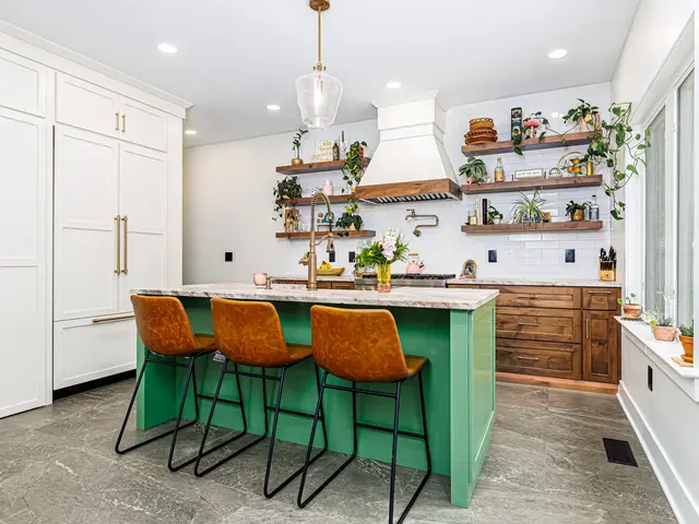 Green kitchen island and woven stools facing wood cabinets, a white range hood and plant-filled floating shelves.