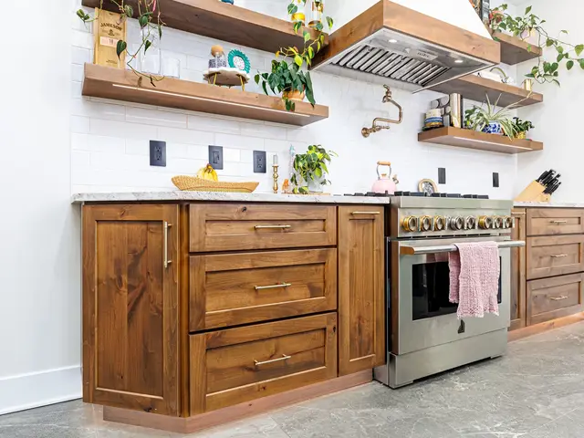 Wood kitchen cabinets and drawers beside a stainless range beneath floating shelves and a wood-trimmed hood.