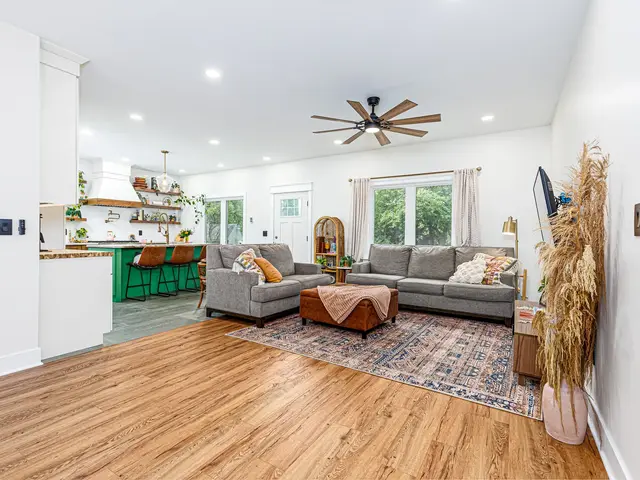 Wide view of the living area and kitchen showing the gray sectional, ceiling fan and green island.