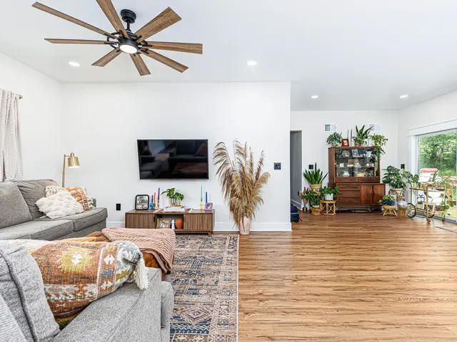 Adjoining living area with a gray sectional, wall-mounted television, ceiling fan and sunlit dining space.